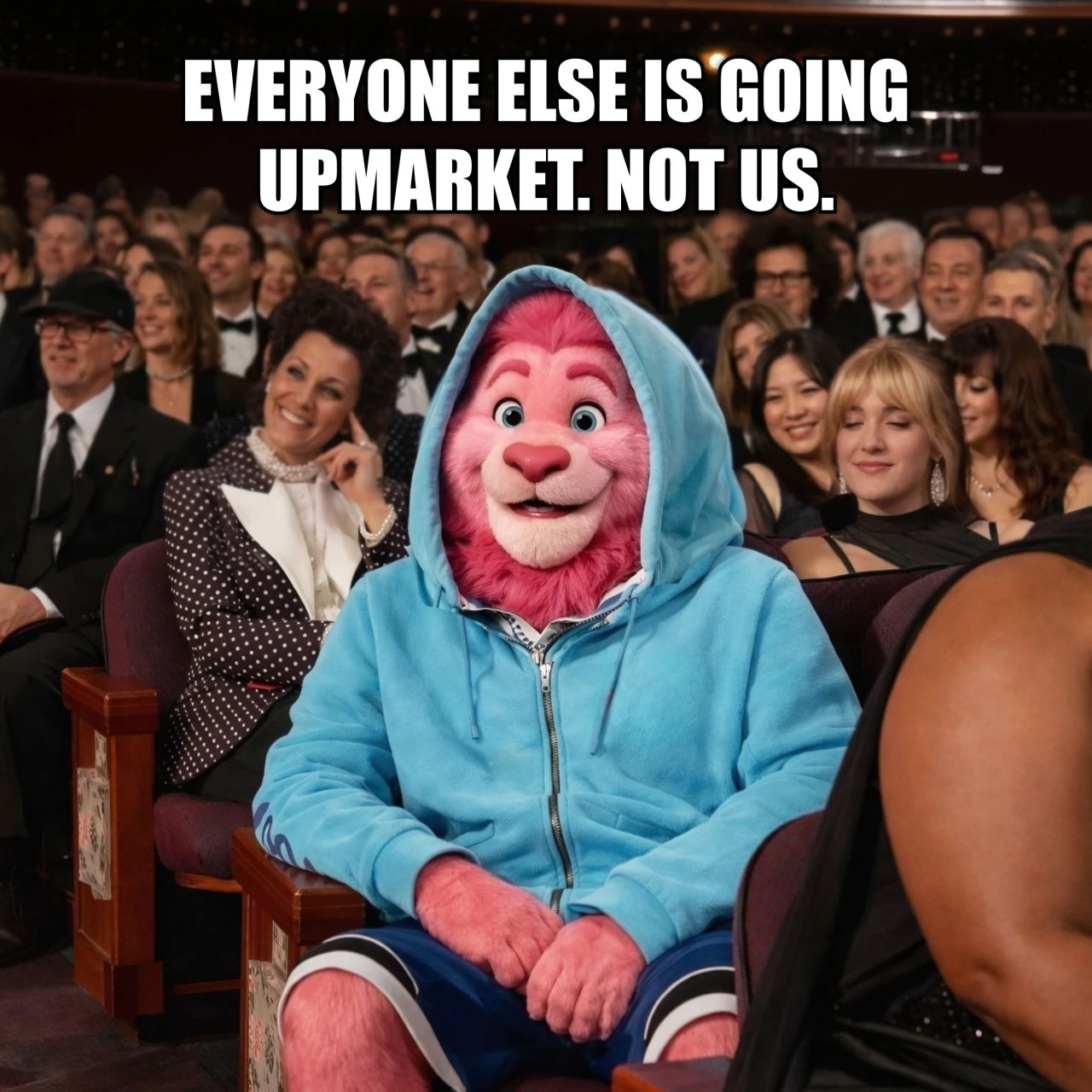 Fibbler meme ad showing their pink lion mascot wearing a casual blue hoodie sitting among formally dressed people at an upscale event. Text reads: "Everyone else is going upmarket. Not us."