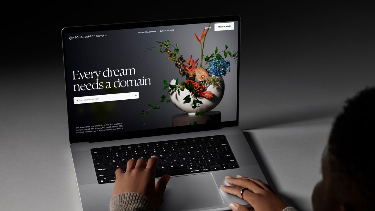 Squarespace Domains ad showing a laptop on a desk displaying a domain search page headlined Every dream needs a domain, with a floral arrangement and a person typing.